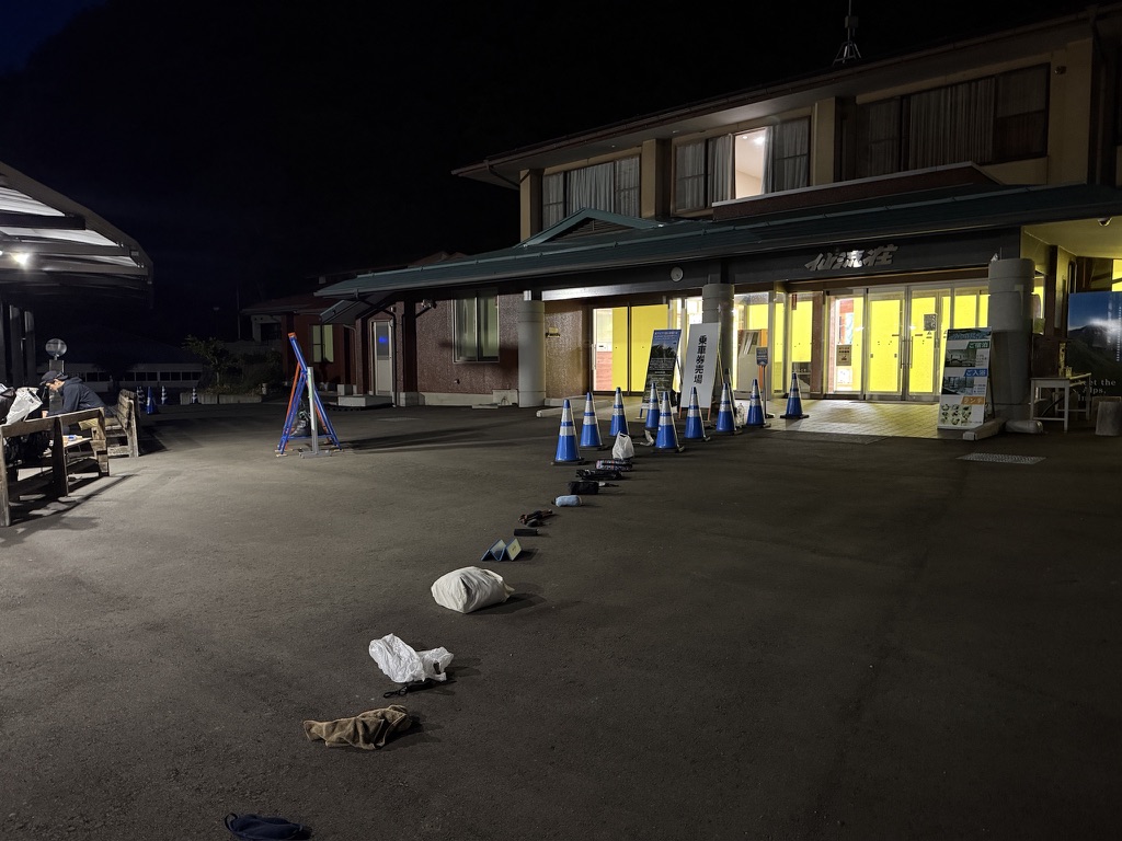 夜の仙流荘前。乗車券売場の看板とコーンに沿って、場所取りの荷物が一列に並んでいる
