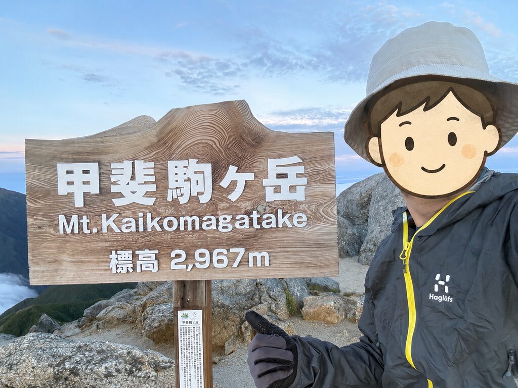 甲斐駒ヶ岳の山頂標識の前に立つ登山者。レインウェアと手袋を着けている
