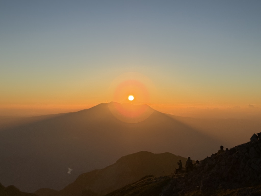 御嶽山の真上に沈んでいく夕日と、稜線で日没を見る登山者のシルエット