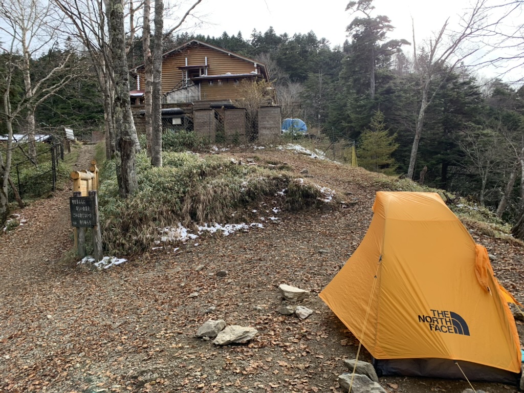 雲取山荘のテント場に張ったマウンテンショット1。落ち葉の地面に雪が残り、奥に木造の山荘が見える