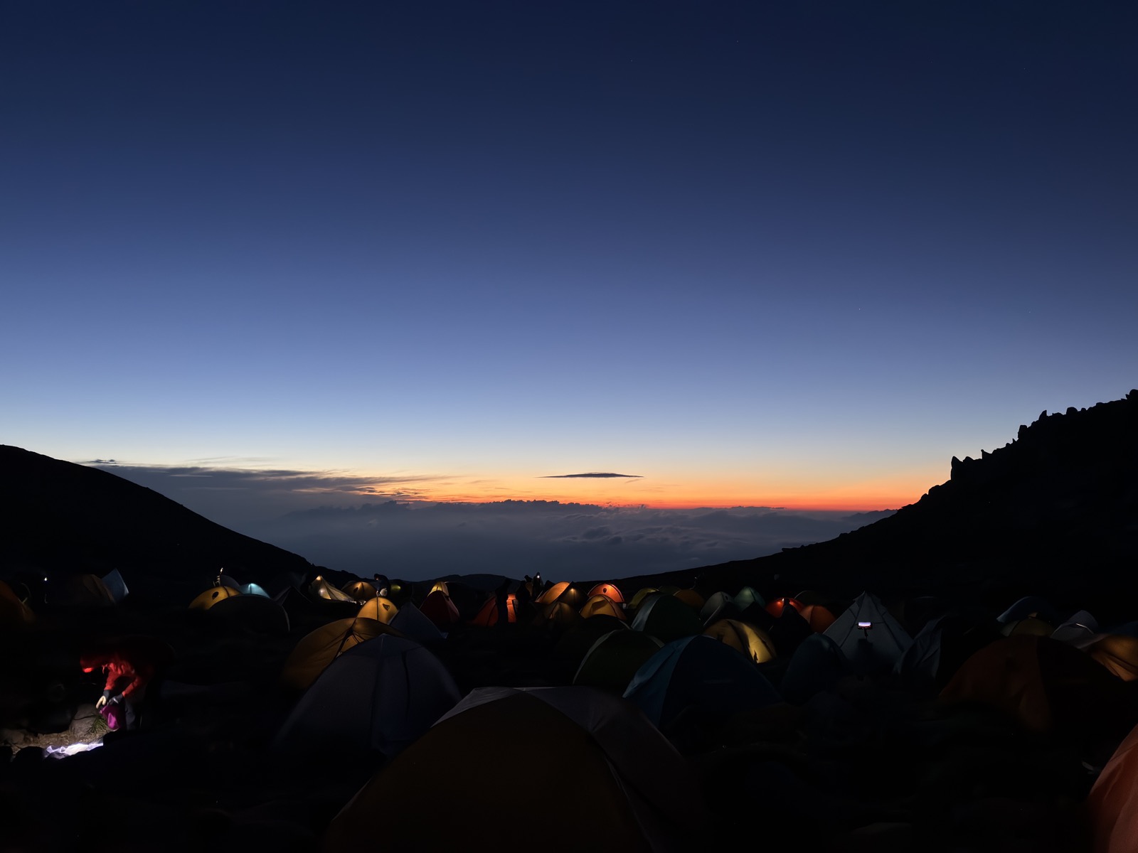 夜明け前のテント場。テントの灯りが点々と灯り、地平線がオレンジに染まりはじめている