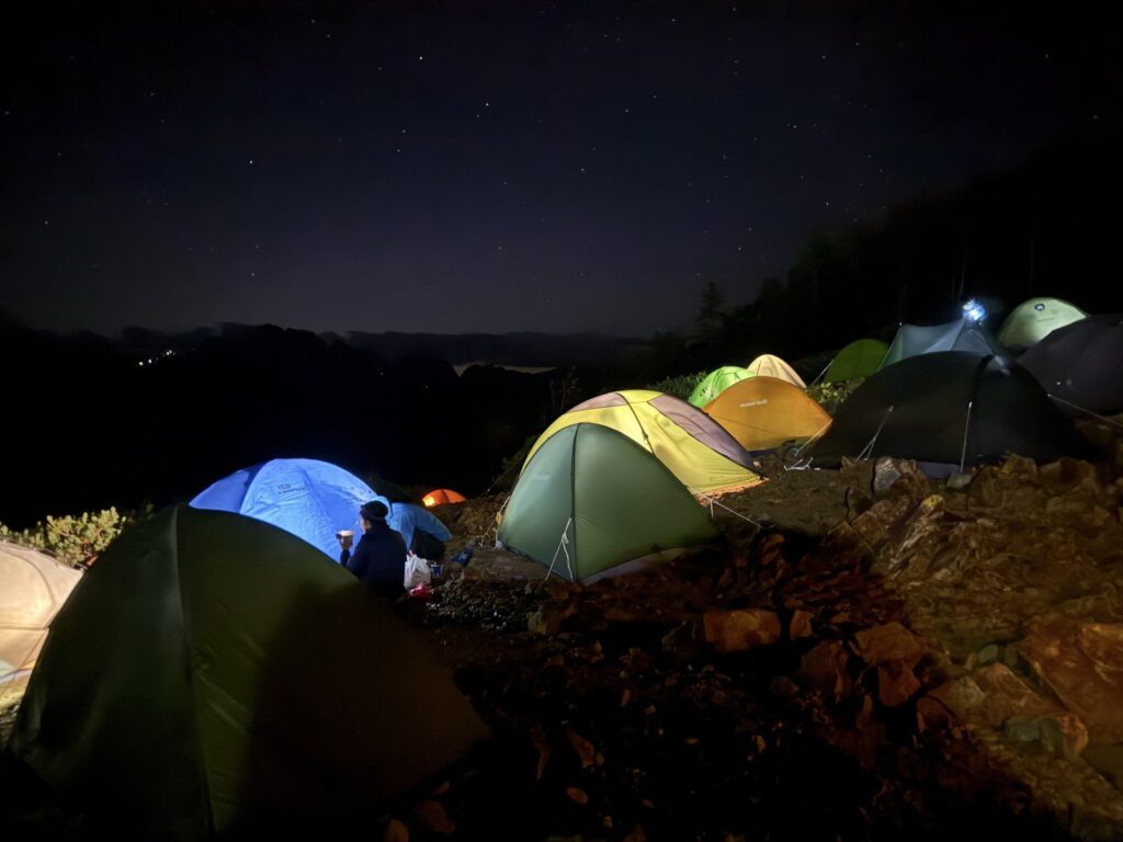 星空の下、明かりのついたテントが並ぶ夜明け前の冷池テント場