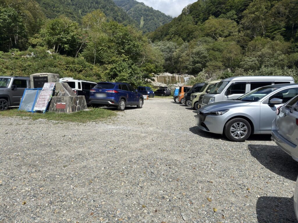 柏原新道登山口そばの砂利の駐車スペース。中部山岳国立公園扇沢の石碑とクマ注意の看板、並んだ車